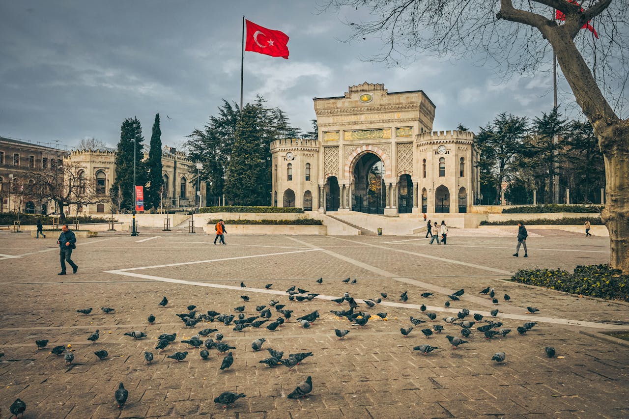 Istanbul University gate with pigeons and Turkish flag in a bustling city square.
