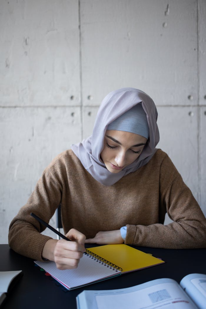 Concentrated Muslim female student in headscarf taking notes in workbook while sitting at table in classroom during lesson at university